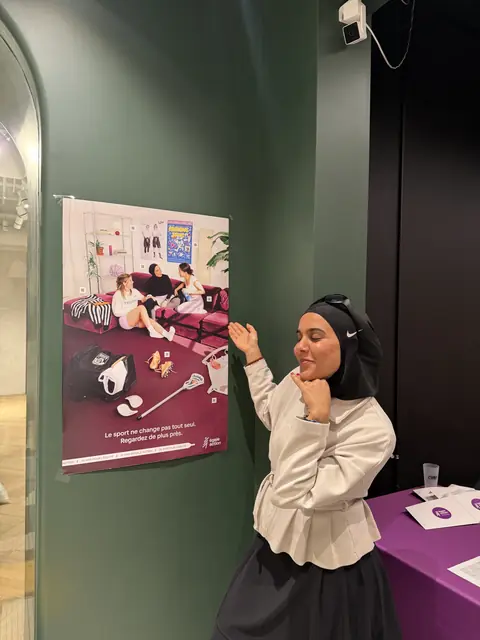 Amani pose à côté d’une affiche de la campagne montrant des femmes voilées en plein effort